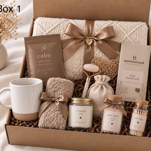 Puede incluir: Una caja de regalo etiquetada como Box 1 que contiene una manta de punto color crema con una cinta marrón, una taza, una bolsa de sales de baño, un rodillo facial de cuarzo rosa, un adorno de corazón, una barra de chocolate con leche Moonstruck, una toalla, una vela, sales de baño y cerillas.