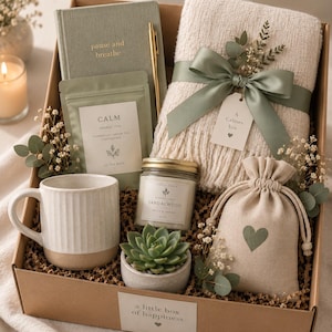 Könnte beinhalten: Eine Geschenkbox mit einem salbeigrünen Notizbuch mit der Aufschrift pause and breathe, einem goldenen Stift, einem salbeigrünen Teebeutel mit der Aufschrift CALM, einer cremefarbenen Strickdecke mit salbeigrünem Band, einer weißen Tasse und einer Kerze.