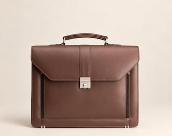 May include: A brown leather briefcase featuring a structured rectangular shape, a top handle, and a central vertical strap with a silver-toned metal push-lock closure. The front includes two vertical zippered pockets on the sides.