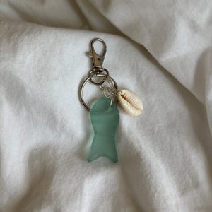May include: A keychain with a silver clasp and ring, featuring a seafoam green glass charm and a small, white seashell. The glass charm is shaped like a stylized fish. The keychain is designed to hold keys or be used as a decorative accessory.