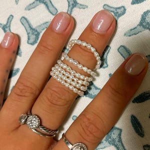 May include: A hand wearing multiple pearl rings stacked on top of each other. The rings are made of small white pearls and are worn on the middle finger. The hand is also wearing a silver ring with a small stone on the ring finger.