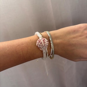 May include: A white beaded bracelet with a pink and white seashell charm. The bracelet is layered with a gold chain bracelet and a gold and black beaded bracelet.