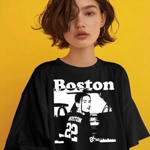 May include: A black short-sleeved t-shirt featuring a white graphic print on the front. The design includes the word Boston in a bold font above a black and white illustration of a person wearing a jersey with the number 22 and the text Boston.