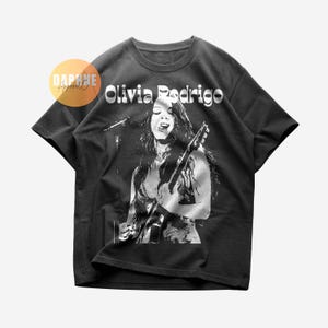 Könnte beinhalten: Ein anthrazitfarbenes Kurzarm-T-Shirt mit einem weißen Grafikdruck von Olivia Rodrigo, die mit Gitarre und Mikrofon auftritt. Der Text Olivia Rodrigo ist in einer fetten, stilisierten Schrift über dem Bild gedruckt. Ein kreisförmiges Logo zeigt Daphne Apparel Co.