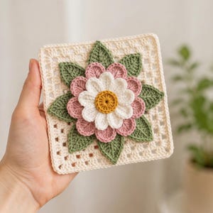 May include: A square crochet granny square featuring a central flower with a yellow center, white inner petals, and dusty pink outer petals. The flower is surrounded by green crocheted leaves on a cream-colored openwork square background.