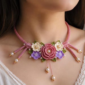 May include: A crochet necklace featuring a central pink rose, flanked by smaller cream and lavender flowers with green leaves. The design includes a pink cord base, accented with white faux pearls and small gold beads, creating a floral collar.