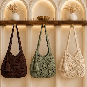 May include: Three crochet shoulder bags hanging in a row, each featuring a square floral motif pattern and a pair of matching tassels on the side. The bags are available in dark brown, sage green, and cream, each with a single strap for carrying.