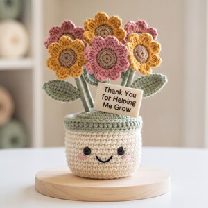 May include: A crocheted potted flower arrangement featuring yellow and pink blossoms with green leaves. The cream-coloured pot has a stitched smiling face with pink cheeks and a rectangular tag that reads Thank You for Helping Me Grow.