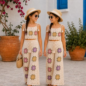 May include: Two matching crochet sets consisting of a cream-coloured crop top and wide-leg trousers, both featuring colourful floral granny square motifs in yellow, pink, and purple. Each set includes a straw hat, sunglasses, and a small woven handbag.