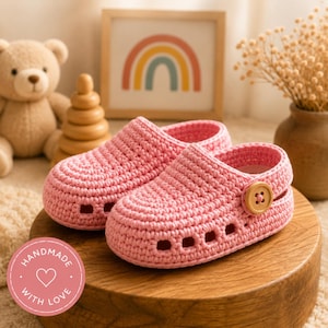 May include: A pair of light pink crochet clogs featuring a rounded toe, side cutouts, and a single wooden button on the outer side of each shoe. The footwear is constructed with a textured yarn stitch pattern and a thick, rounded sole design.