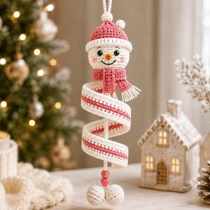 May include: A crocheted snowman ornament featuring a pink hat and scarf, with a long, spiralling white and pink body. The snowman has a small orange nose, black eyes, and a smiling mouth. Two small pink beads and two white spheres hang at the base.