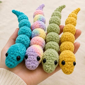 May include: Four small, segmented crochet snakes with smooth, plush-like texture. The snakes are colored in bright turquoise, a pastel rainbow gradient, light sage green, and solid yellow. Each snake features two small, round, black plastic eyes.