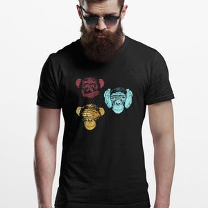 Puede incluir: Una camiseta negra de manga corta con un estampado gráfico de tres monos. Un mono rojo se cubre la boca, uno amarillo se cubre los ojos y uno azul claro se cubre las orejas, dispuestos en un patrón triangular en la prenda.
