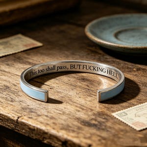 Puede incluir: Una pulsera tipo brazalete de tono plateado con una incrustación de esmalte azul claro en el exterior. La superficie interior está grabada con el texto negro "This too shall pass, BUT FUCKING HELL" en una fuente serif clara y abierta.