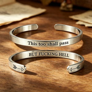 May include: Two silver-toned metal cuff bracelets with open ends. The top bracelet is engraved with the text "This too shall pass". The bottom bracelet is engraved with the text "BUT FUCKING HELL" and features small arrow icons at both ends.