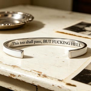 May include: A silver-toned cuff bracelet featuring a white inlay along the outer band. The interior surface is engraved with the black text, "This too shall pass, BUT FUCKING HELL". The bracelet has an open design with rounded ends.