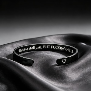 May include: A black metal cuff bracelet featuring the engraved text "This too shall pass, BUT FUCKING HELL" along the inner band and a small white heart symbol engraved on the outer surface near the open end of the bracelet.