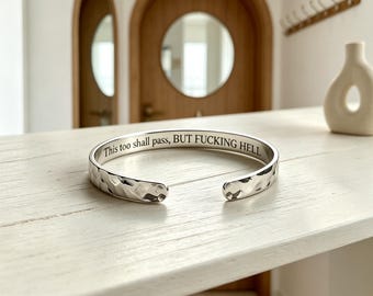 May include: A silver-toned metal cuff bracelet with a hammered, textured exterior surface. The interior of the bracelet features the engraved black text, "This too shall pass, BUT FUCKING HELL," written in a clear, serif font along the inner band.