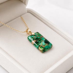 May include: A gold chain necklace featuring a rectangular pendant with a green and copper-coloured marbled stone inlay. The pendant has a polished finish and is suspended from a delicate gold-tone metal chain, presented inside a white box.