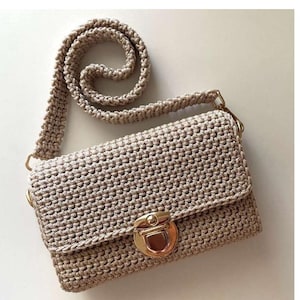 May include: A beige crochet crossbody bag featuring a textured, knotted stitch pattern. The rectangular bag has a flap closure secured with a polished gold-tone metal push-lock clasp and includes a matching thick, braided crochet shoulder strap.