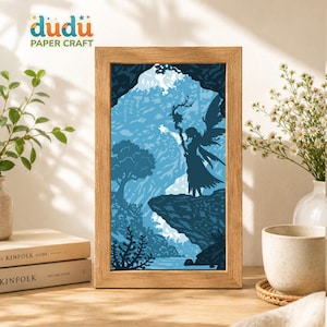 May include: A rectangular wooden frame containing a layered paper craft art piece. The artwork depicts a silhouette of a winged figure holding a staff, standing on a cliff edge overlooking a blue-toned forest landscape with trees and water.