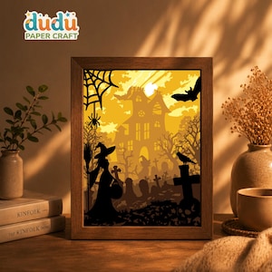May include: A rectangular wooden frame containing a layered paper craft scene with a backlit yellow glow. The silhouette design features a witch, a haunted house, a graveyard with tombstones, a spider web, a bat, and a crow against a bright sky.