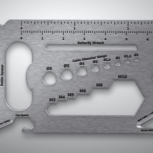 May include: A rectangular silver-colored metal multi-tool featuring a ruler with inch and centimeter markings, a bottle opener, a can opener, a box cutter, a butterfly wrench, a cable diameter gauge, a wire cutter, a nail puller, and a screwdriver.