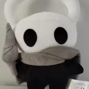 May include: A plush toy character featuring a white head with two curved horns, large black circular eyes, and a grey cloak draped over a black body. The character holds a grey pointed weapon in its right hand and has a white tag attached.