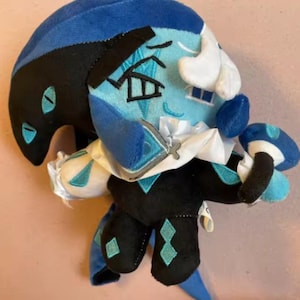 Pode incluir: Um peluche representando uma personagem com a cabeça dividida em duas cores, um lado preto e outro azul claro. A figura tem um colarinho branco com folhos, um corpo preto com detalhes em losango azul claro e segura um pequeno objeto em forma de cogumelo.