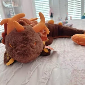 Puede incluir: Un dragón de peluche con pelaje marrón oscuro, cuernos naranjas y detalles naranjas a lo largo de la espalda y la cola. La criatura tiene ojos naranjas bordados y un cuerpo largo y segmentado. Una pequeña etiqueta está unida al costado del peluche.