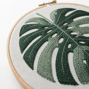 May include: A circular wooden embroidery hoop framing a piece of light beige fabric. The fabric features a hand-stitched monstera leaf design rendered in various shades of dark and light green thread, showcasing detailed leaf veins and cutouts.