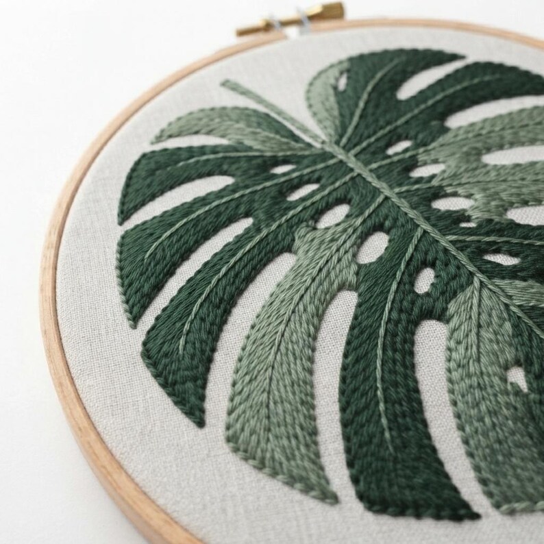 May include: A circular wooden embroidery hoop framing a piece of light beige fabric. The fabric features a hand-stitched monstera leaf design rendered in various shades of dark and light green thread, showcasing detailed leaf veins and cutouts.