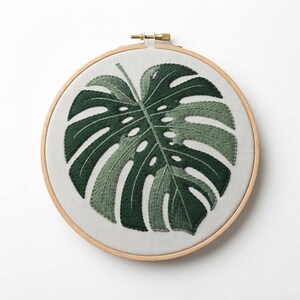 May include: A circular wooden embroidery hoop containing a piece of light gray fabric. The fabric features a hand-stitched monstera leaf design rendered in various shades of dark and forest green thread, showcasing detailed leaf fenestrations.