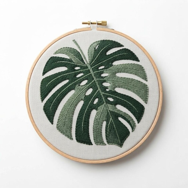May include: A circular wooden embroidery hoop containing a piece of light gray fabric. The fabric features a hand-stitched monstera leaf design rendered in various shades of dark and forest green thread, showcasing detailed leaf fenestrations.