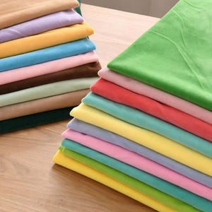 Può includere: Due pile di tessuti piegati in vari colori a tinta unita, tra cui verde, rosa, rosso, giallo, blu, marrone e beige. I pezzi di tessuto sono sovrapposti ordinatamente, mostrando una consistenza liscia e una varietà di tonalità vivaci.