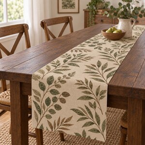 May include: A rectangular table runner made of light beige fabric featuring a printed pattern of green and brown leaves. The runner is laid flat across a dark brown wooden dining table, extending from one end to the other with a botanical design.