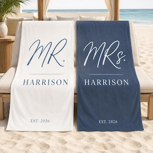 May include: Two rectangular beach towels, one white with dark blue text and one dark blue with white text. Both feature the script text MR. or MRS. above the name HARRISON and the text EST. 2026 in a clean, sans-serif font at the bottom.