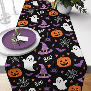 Könnte beinhalten: Ein schwarzer Tischläufer mit einem sich wiederholenden Muster aus orangefarbenen Kürbissen, weißen Geistern, violetten Hexenhüten, weißen Spinnennetzen, kleinen violetten Spinnen und dem Wort Boo in weißer Schrift, akzentuiert durch kleine orange und rosa Sterne.