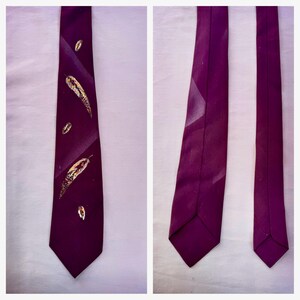 May include: A deep purple necktie featuring a series of hand-painted feathers in shades of gold, brown, and white. The design is arranged vertically along the front blade of the tie, while the back view shows the plain solid purple fabric.