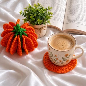以下が含まれることがあります： 緑色の茎を持つかぎ針編みのオレンジ色のカボチャ、コーヒーが入った陶器のマグカップ、そして丸いオレンジ色のかぎ針編みのコースター。マグカップにはオレンジ色の花と緑の葉の模様が描かれています。カボチャの後ろには小さな緑の植物が置かれています。