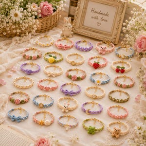 May include: A collection of handmade crochet bracelets featuring various charms and patterns, including flowers, hearts, bows, stars, shells, moons, and small animal figures like bears, rabbits, and frogs, arranged on a light fabric surface.