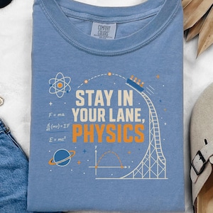 May include: A light blue t-shirt featuring a graphic with the text STAY IN YOUR LANE, PHYSICS, an atom, a roller coaster, a planet, and physics equations. The shirt is displayed alongside blue denim shorts, black sunglasses, and a gold chain necklace.