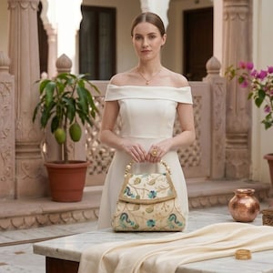 Può includere: Un abito color crema con spalle scoperte abbinato a una borsa strutturata caratterizzata da un intricato ricamo floreale nei toni del blu, verde e oro, accentuato da un manico superiore decorato con perle sferiche bianche.