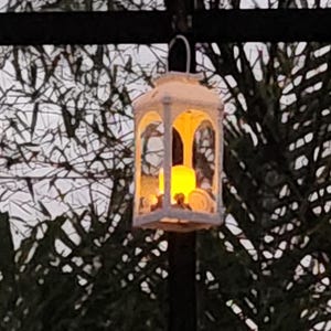 May include: A white rectangular lantern with a metal frame and glass panels hanging from a dark support. Inside the lantern, a glowing yellow LED candle emits a warm light, illuminating the interior space and casting a soft glow through the glass.
