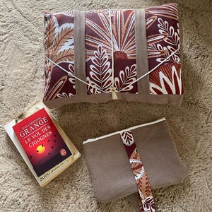 Peut inclure: Un coussin rectangulaire à motif botanique bordeaux et blanc, doté de deux sangles en tissu beige et d'un cordon blanc. À côté, une pochette beige plus petite avec une sangle à motif assorti et le livre intitulé Le Vol des Cigognes.