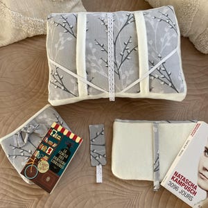 Peut inclure: Un coussin rectangulaire gris à motif de branches de saule, avec des bandes de tissu blanc et une bordure en dentelle centrale. L'ensemble comprend une housse de livre grise assortie, un petit marque-page et une couverture de livre crème.