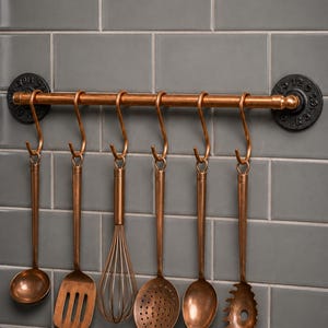 Puede incluir: Un soporte para utensilios de cocina en tono cobre montado con bridas circulares negras de estilo industrial. Seis utensilios a juego cuelgan de ganchos en forma de S, incluyendo un cucharón, una espátula, un batidor, una espumadera y un servidor de pasta.