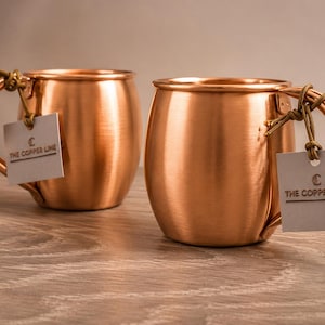 May include: Two polished copper mugs with rounded bodies and curved handles. Each mug features a small, square, light-coloured paper tag tied to the handle with a thin cord. The tags display the text THE COPPER LINE with a small logo above it.