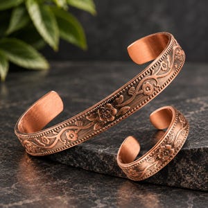 May include: A copper cuff bracelet and a matching copper ring, both featuring an embossed floral vine pattern with dark oxidised detailing. The metal has a warm, reddish-brown tone with a polished finish on the inner bands and textured edges.