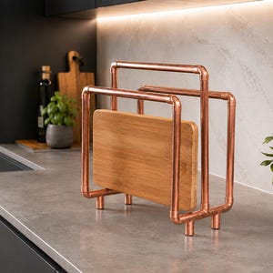 May include: A copper pipe rack designed to hold cutting boards, featuring a rectangular frame constructed from metallic copper-coloured pipes and fittings, with a single rectangular wooden cutting board positioned inside the central section.